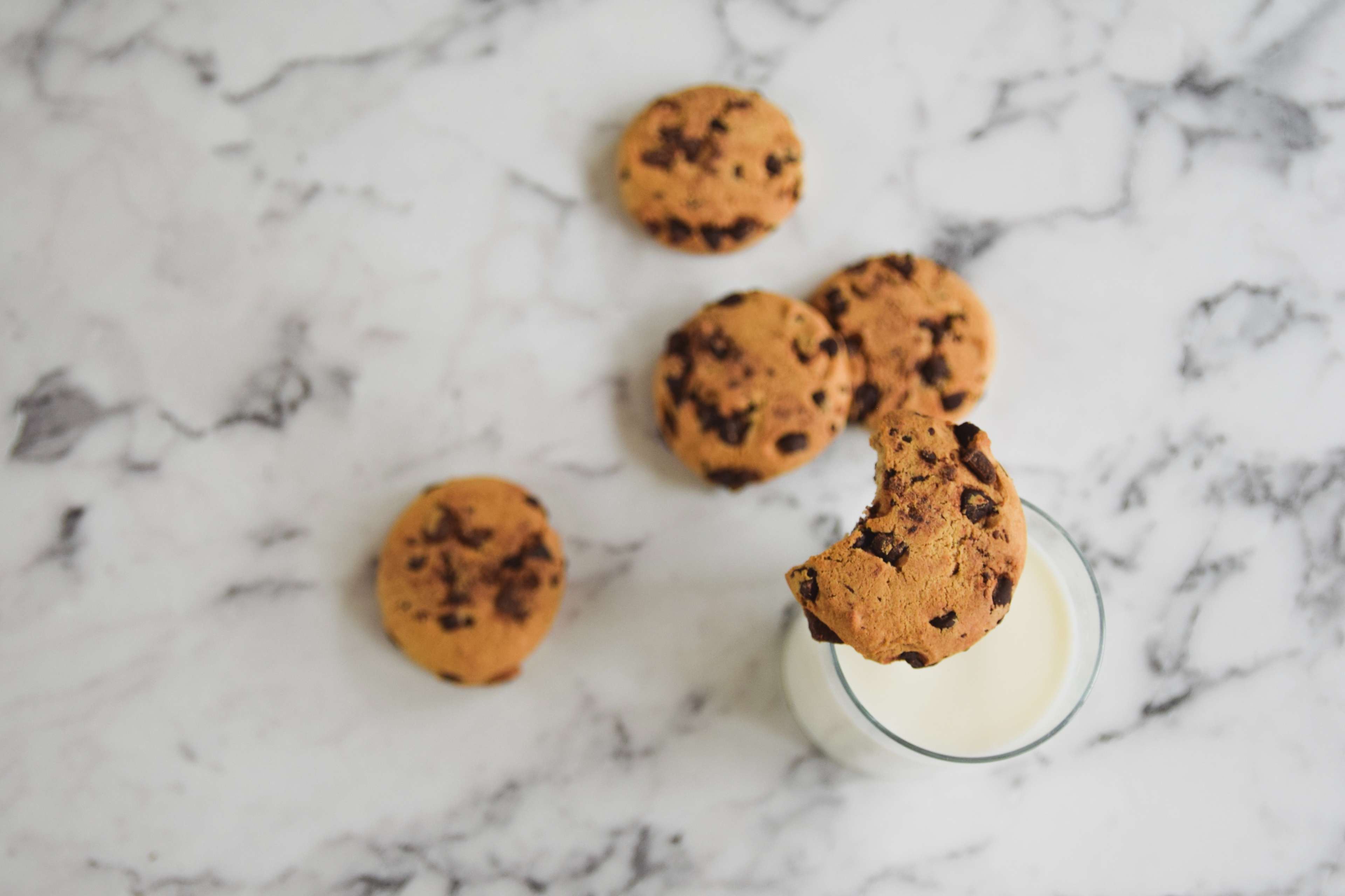 5 chocolate chip cookies on top a quartz countertop with one on top a glass of milk with a bite taken out