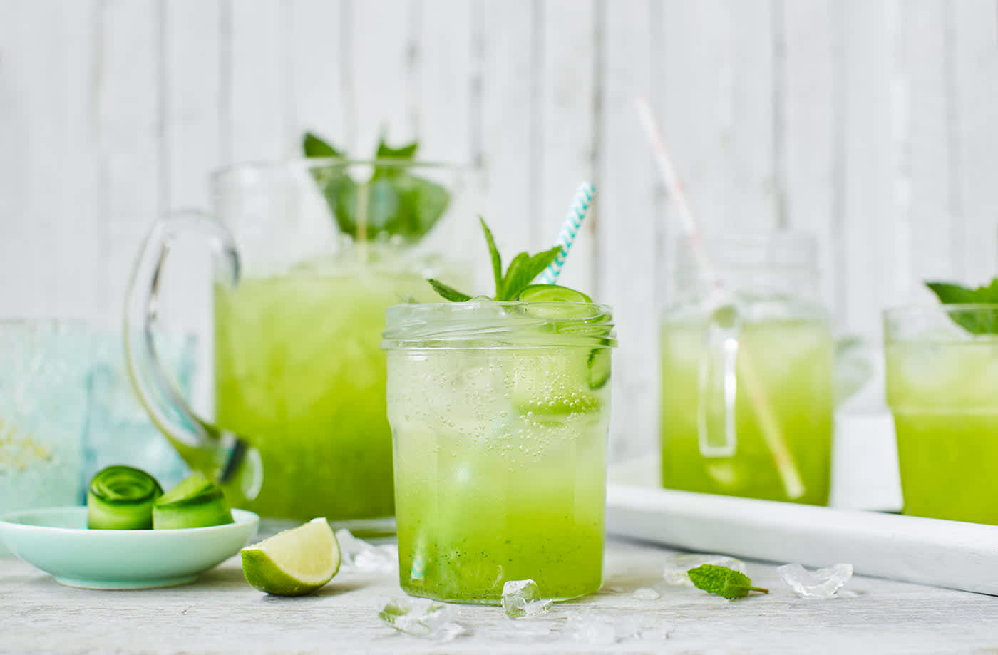 Glass & pitcher full of cucumber, lime & elderflower cooler garnished with mint and lime