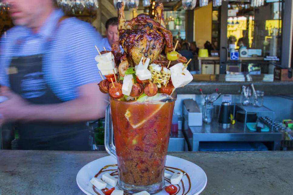 A Bloody Mary cocktail in a large glass pitcher garnished with tomatoes, olives, mozzarella cheese and a rotisserie chicken. 