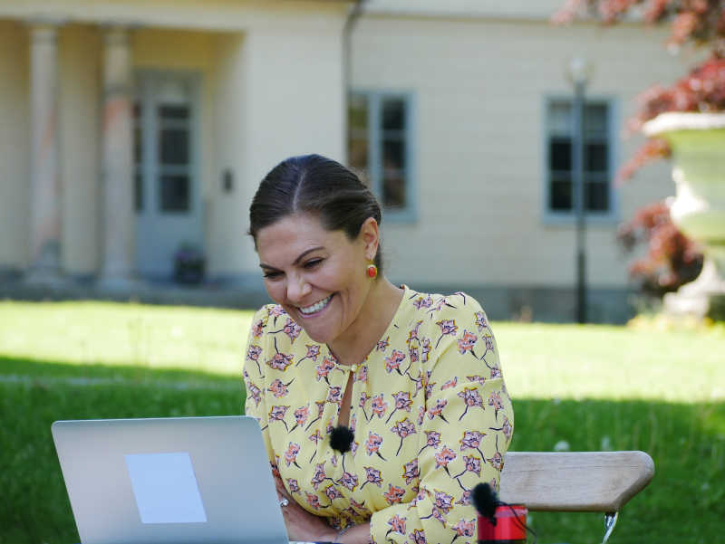 Kronprinsessan Victoria sitter vid en bord med en uppslagen bärbar dator framför sig, hon ler brett mot datorn.