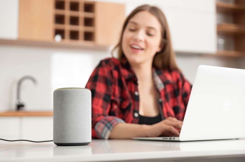 A woman sitting at a table, using a laptop and a smart speaker for work and communication.