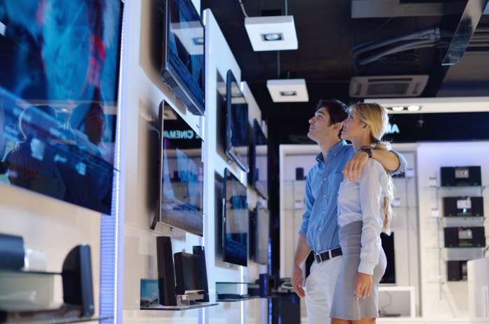 A couple stands closely together in an electronics store, looking at a wall display of flat-screen televisions