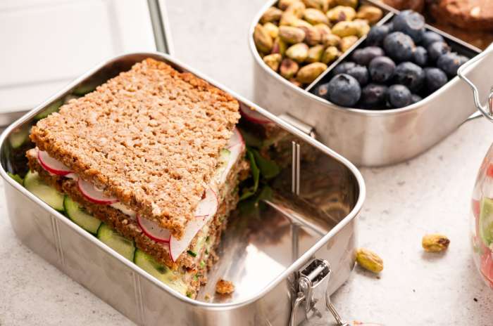 A wholesome sandwich with vegetables in a metal lunchbox, next to a container of pistachios and blueberries