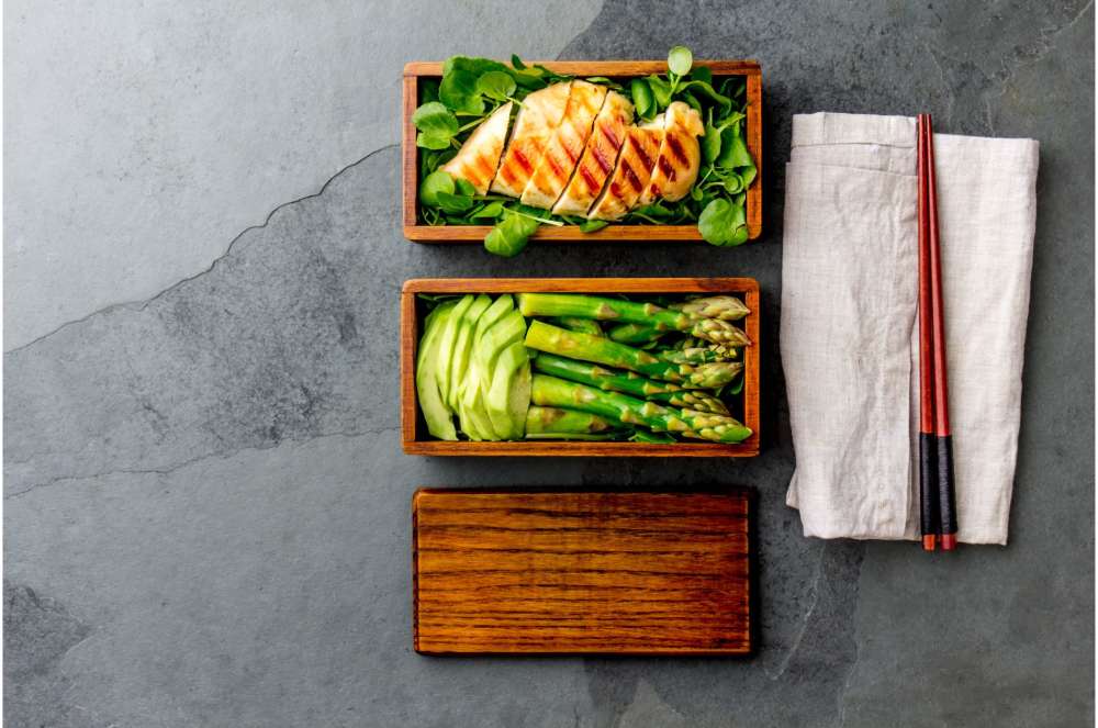 Asparagus, chicken, and rice in wooden bento boxes on stone background