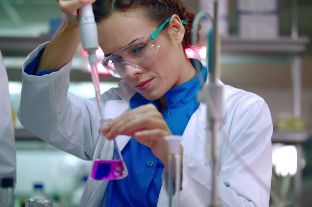 A lab testing professional in a coat and blue gloves using a pipette to transfer a purple liquid into a test tube