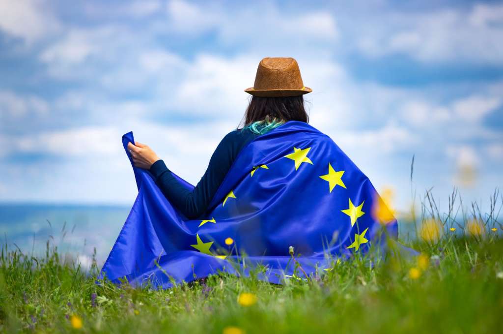 A person sitting in a grassy field wrapped in a European Union flag with a brown hat on their head looking at a cloudy sky.