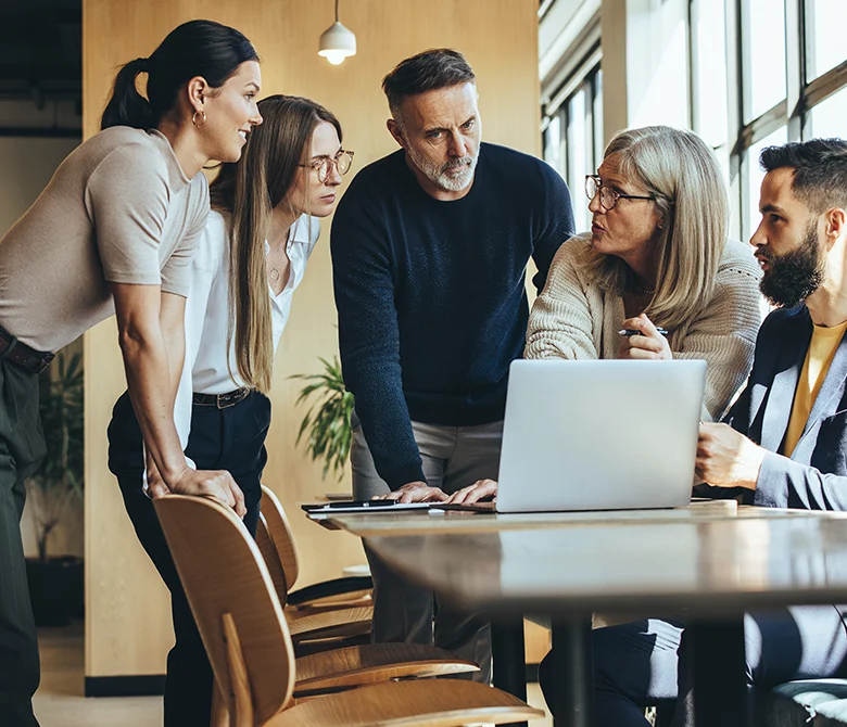 Group of colleagues, 3 females and two males, gathered around laptop in discussion.