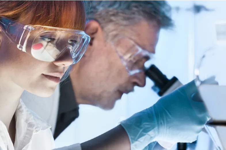 Two medical researchers wearing goggles looking into microscopes