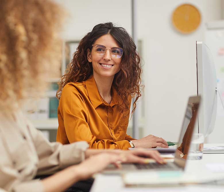 Young woman with curly long hair and glasses sitting at her desk, smiling off camera at a coworker