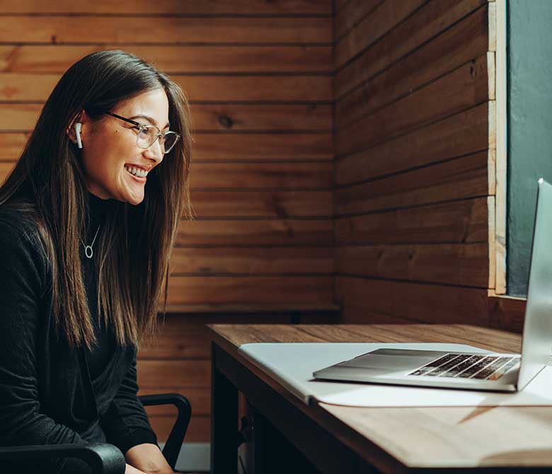 Young woman with long brown hair and glasses sitting at computer smiling, on a video call.
