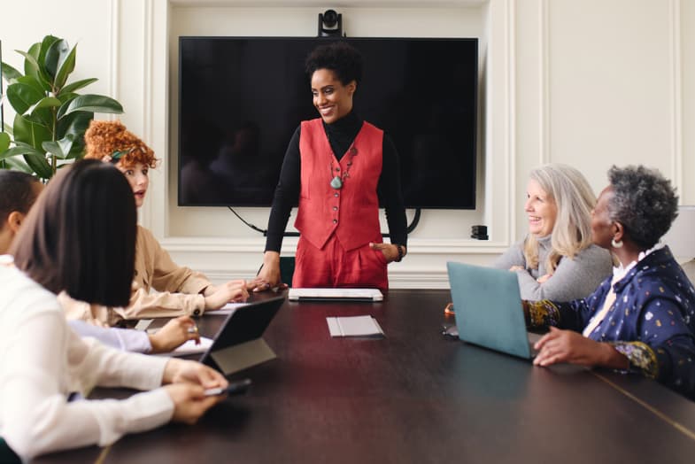 Coworkers in casual office setting meeting room sitting around a conference table looking up at woman standing at head of the table smiling.