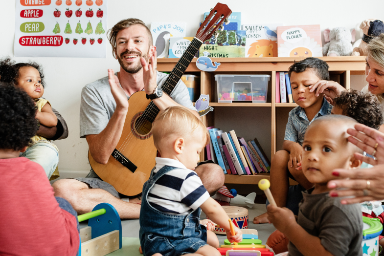 Young male daycare worker playing guitar, sitting and singing in circle with students 