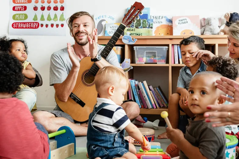 Young male daycare worker playing guitar, sitting and singing in circle with students