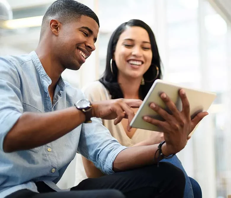 Young man sitting next to young woman colleague, showing her information on his tablet device.
