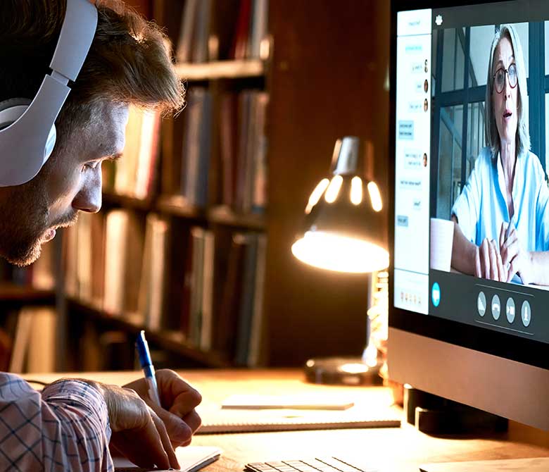 Man sitting at desktop computer attending a virtual workshop, writing notes and wearing headphones.