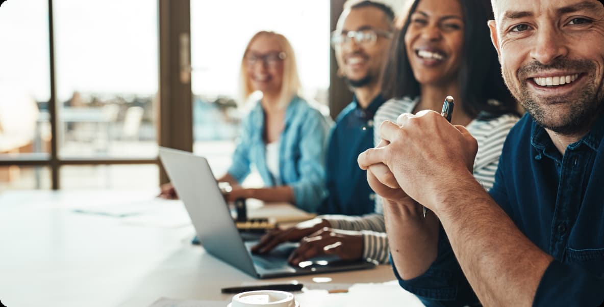 young male office worker sitting with 3 other colleagues behind him blurred around conference table smiling at camera