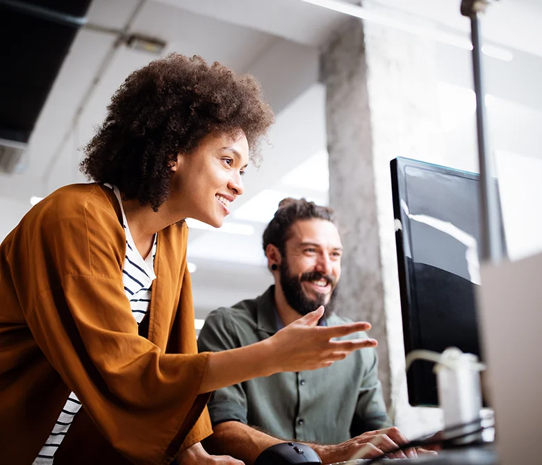 Woman standing next to male coworker sitting down working at laptop.