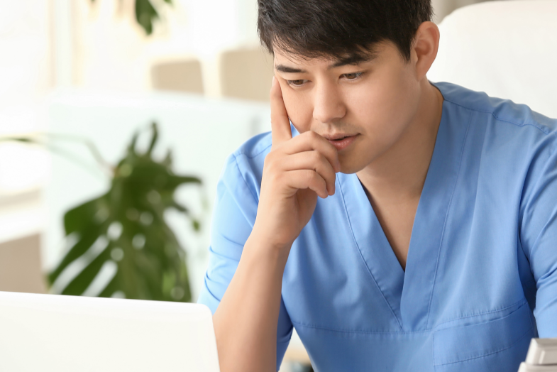 Young man in scrubs looking deep in thought staring at laptop 