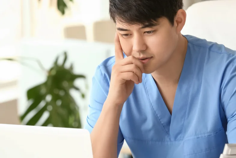 Young man in scrubs looking deep in thought staring at laptop