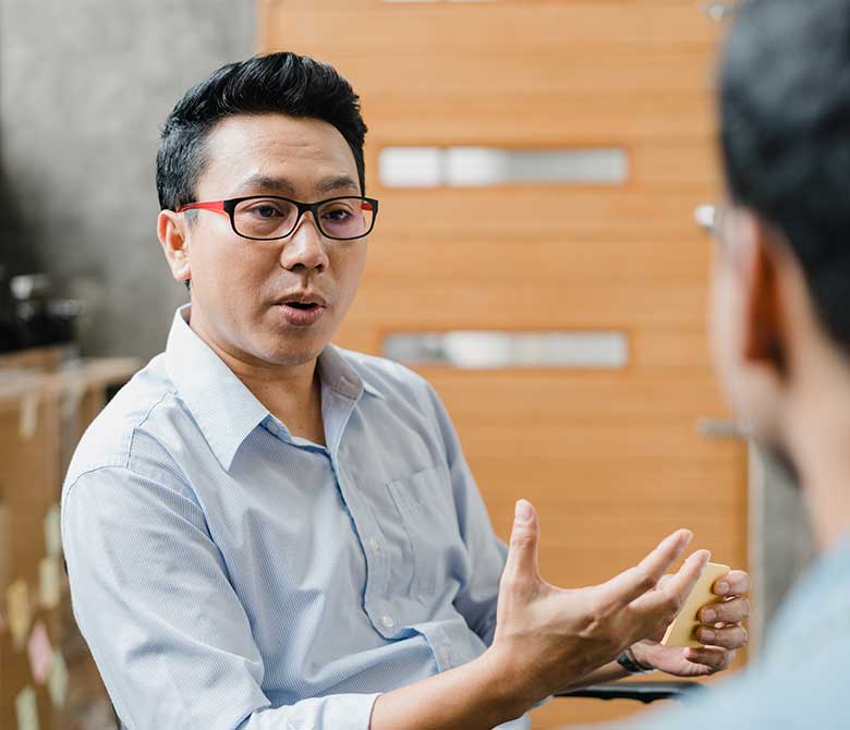 Young asian man with glasses talking to colleague who is not facing the camera, in mid discussion.