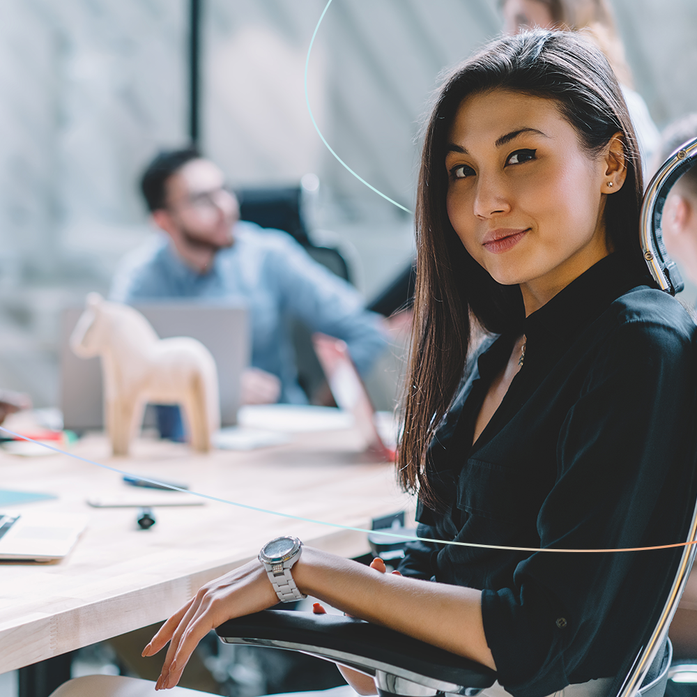 Young woman sitting at office desk, turning to look at camera with soft smile.