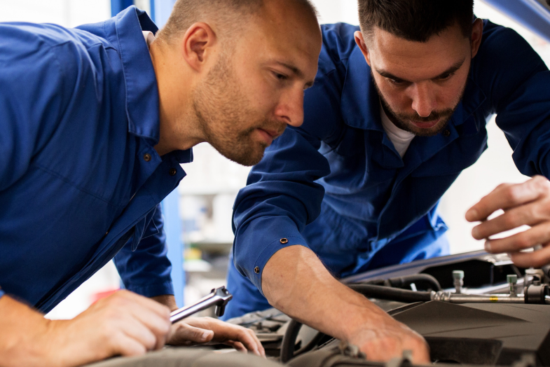 two men in jumpsuits working under hood of car