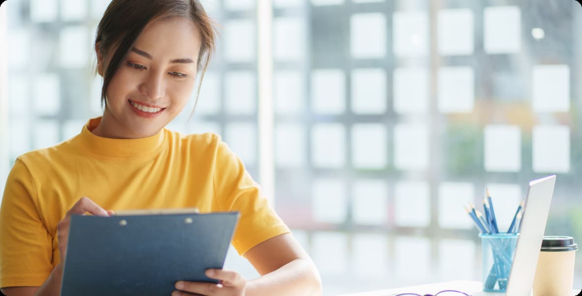 young woman in casual professional attire sitting at a desk smiling holding a clipboard