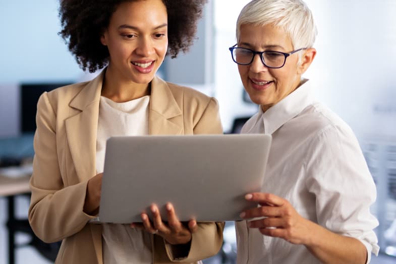 young woman standing with older woman, holding laptop smiling, both in office attire