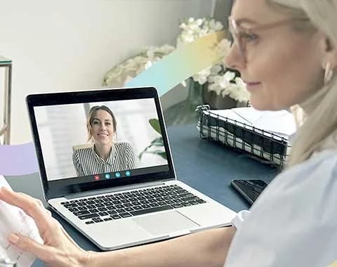 Older woman interviewing young woman on laptop screen while looking at printed resume.