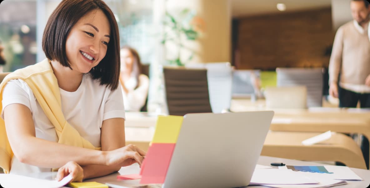 young asian woman in open office setting sitting smiling at laptop