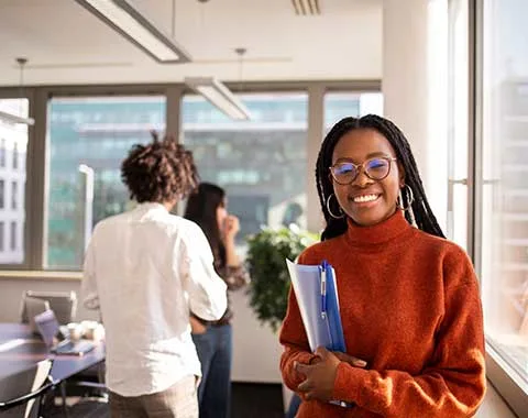 Young woman wearing glasses in office setting smiling holding reports and looking at the camera.