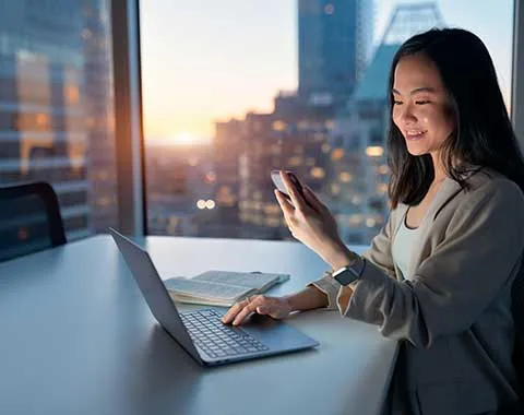Young female office worker smiling, looking at phone while working on laptop