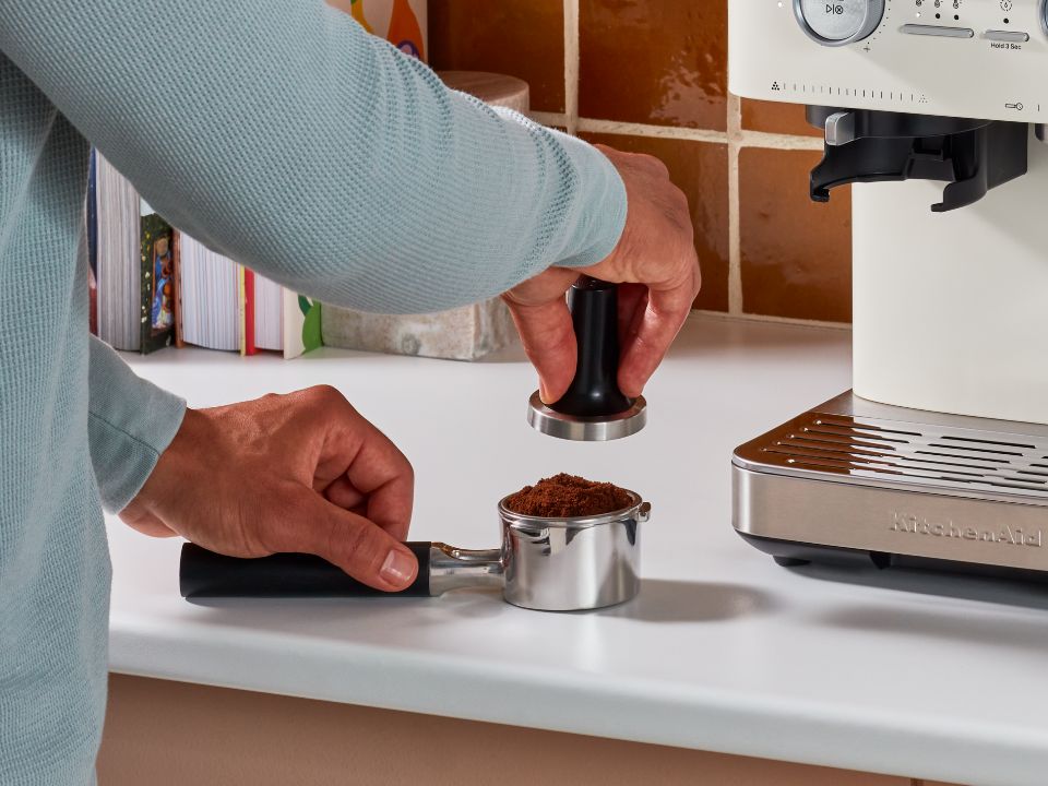 Semi-automatic-espresso-machine-with-burr-grinder-5KES6551-person-using-tamper-to-press-the-ground-coffee-in-portafilter-next-to-procelain-coffee-machin