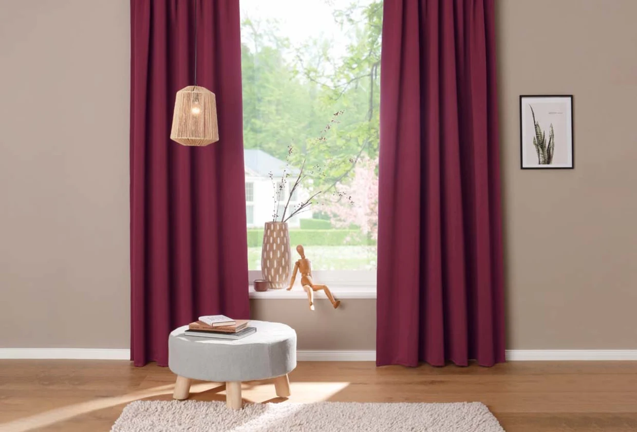 Wohnzimmer mit bodenlangen weinroten Vorhängen, grauem Hocker, Vase auf der Fensterbank und einer hängenden Naturfaserlampe. Wohnzimmer mit bodenlangen weinroten Vorhängen, grauem Hocker, Vase auf der Fensterbank und einer hängenden Naturfaserlampe.