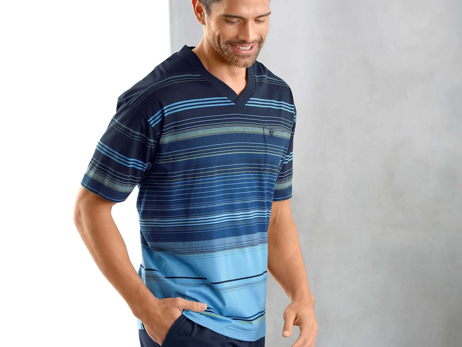 Ein männliches Model trägt ein kurzärmeliges Shirt mit blauem Farbverlauf und Streifenmuster sowie eine dunkle Hose, steht vor hellem Hintergrund.