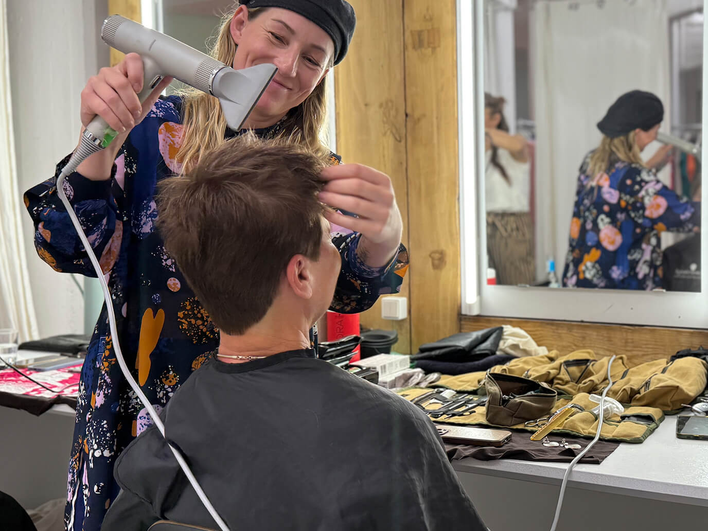 Das Bild zeigt eine Szene aus dem Backstage-Bereich: Eine Stylistin föhnt einer Frau die Haare vor einem Spiegel, auf dem Tisch liegen verschiedene Stylingprodukte, Bürsten und Handtücher, alles in einer freundlichen und entspannten Atmosphäre.