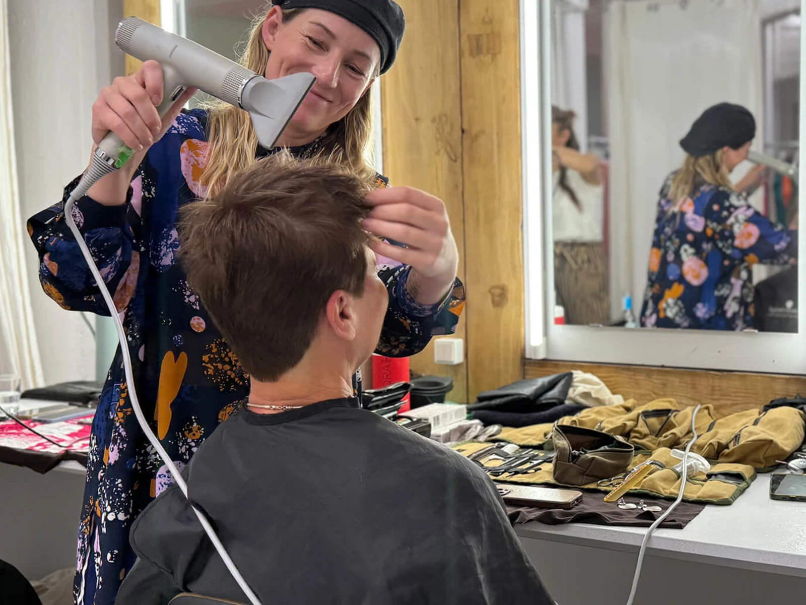 Das Bild zeigt eine Szene aus dem Backstage-Bereich: Eine Stylistin föhnt einer Frau die Haare vor einem Spiegel, auf dem Tisch liegen verschiedene Stylingprodukte, Bürsten und Handtücher, alles in einer freundlichen und entspannten Atmosphäre. Das Bild zeigt eine Szene aus dem Backstage-Bereich: Eine Stylistin föhnt einer Frau die Haare vor einem Spiegel, auf dem Tisch liegen verschiedene Stylingprodukte, Bürsten und Handtücher, alles in einer freundlichen und entspannten Atmosphäre.