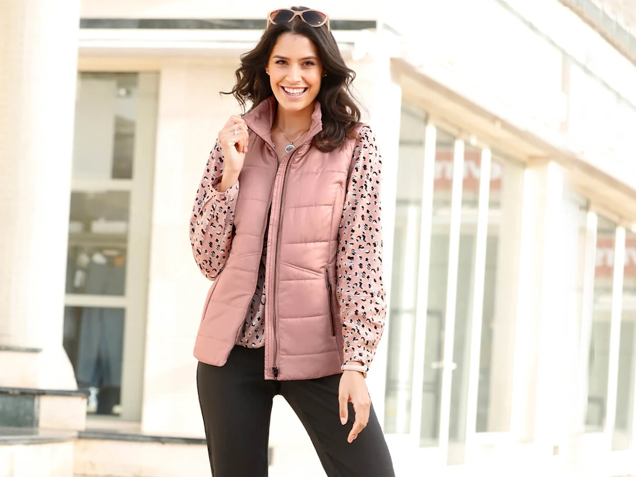 Dame trägt eine rosafarbene Steppweste über einer gemusterten Bluse und einer schwarzen Hose, stehend vor einem hellen Gebäude mit Glasfassade. Dame trägt eine rosafarbene Steppweste über einer gemusterten Bluse und einer schwarzen Hose, stehend vor einem hellen Gebäude mit Glasfassade.