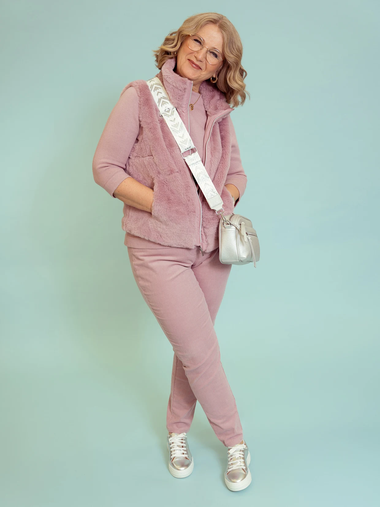 Eine lächelnde Frau mit Brille und welligem blondem Haar trägt eine rosa Kunstfellweste über einem passenden rosa Oberteil und einer Hose. Sie hat eine silberne Umhängetasche mit gemustertem Riemen und silber-weiße Sneaker an. Sie steht mit gekreuzten Beinen vor einem hellblauen Hintergrund.