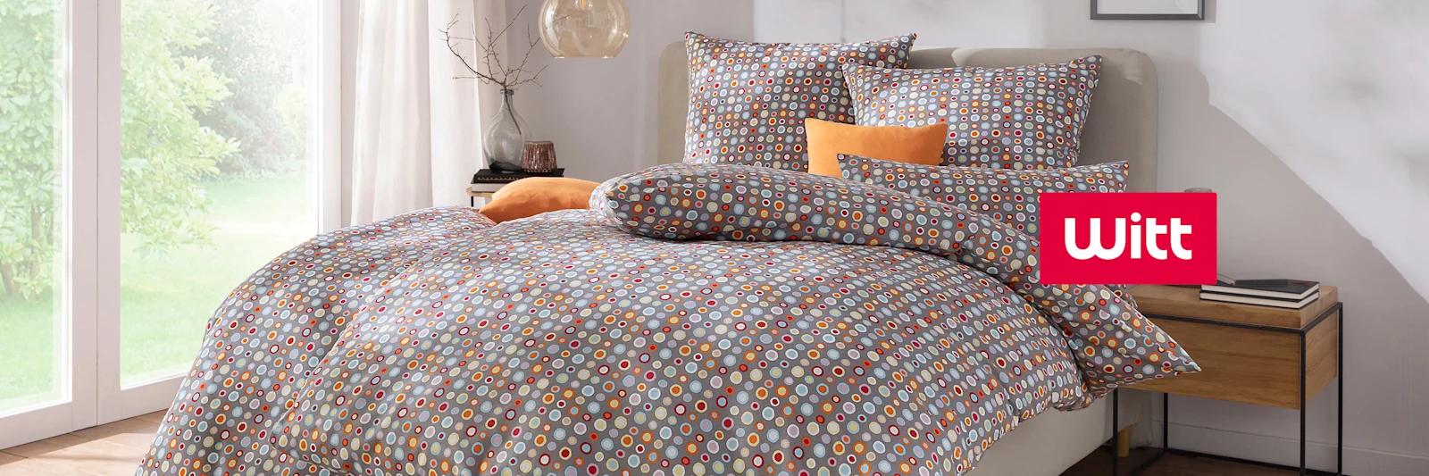 Ein Bett mit bunter Bettwäsche in Grau, Orange und Weiß, dekoriert mit mehreren Kissen, steht in einem hellen Schlafzimmer mit großem Fenster. Ein Bett mit bunter Bettwäsche in Grau, Orange und Weiß, dekoriert mit mehreren Kissen, steht in einem hellen Schlafzimmer mit großem Fenster.