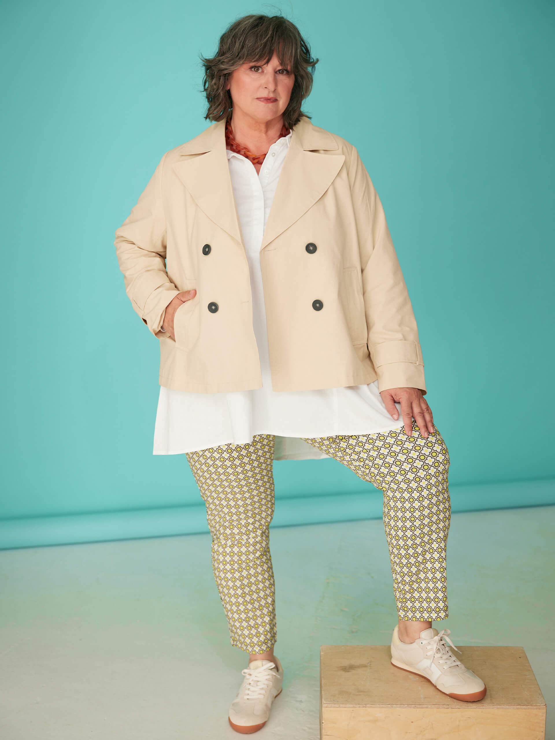 Ein Model trägt eine beigefarbene Jacke, weiße Bluse, gemusterte Hose und Sneaker vor türkisfarbenem Hintergrund.
