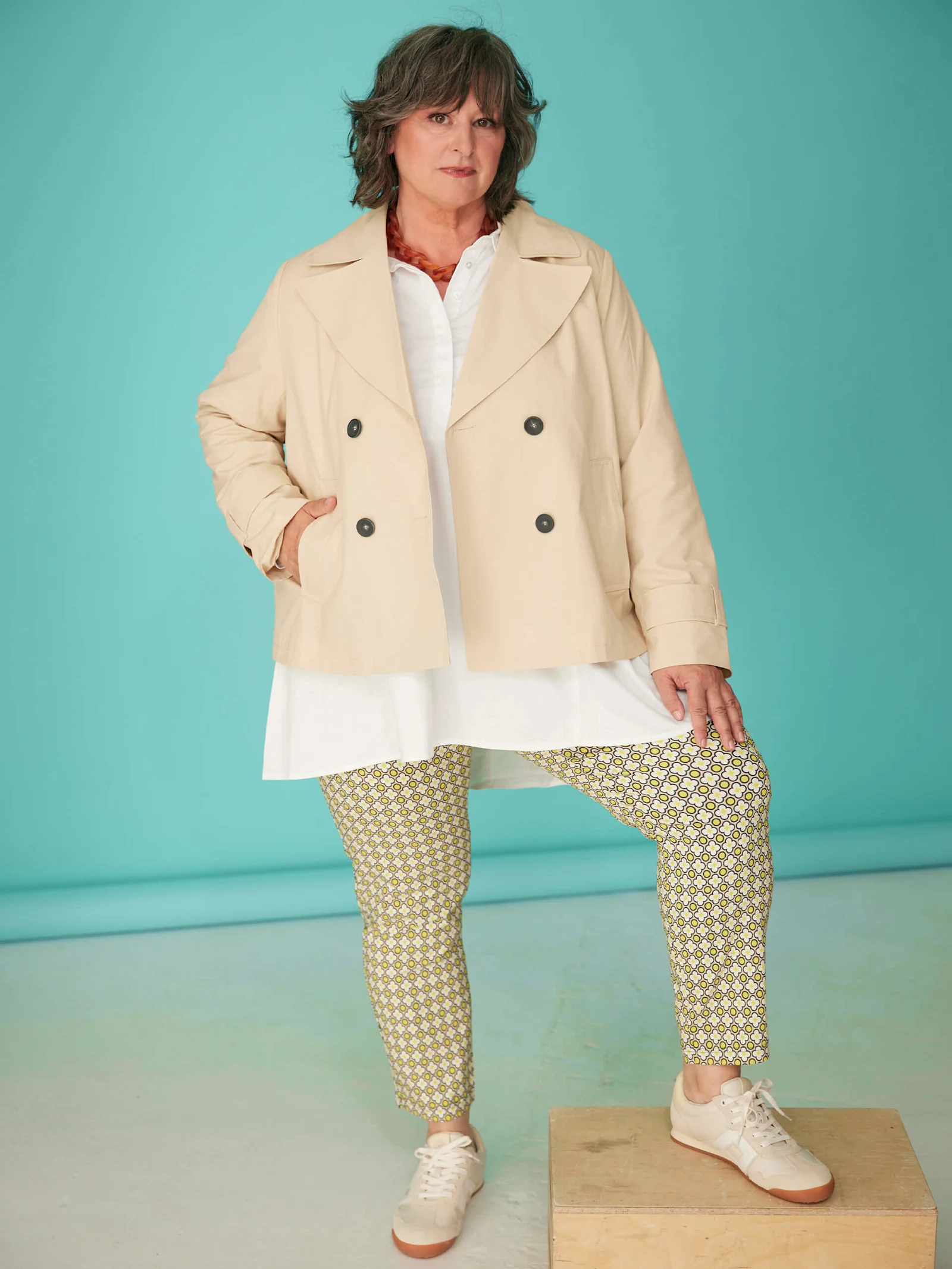 Ein Model trägt eine beigefarbene Jacke, weiße Bluse, gemusterte Hose und Sneaker vor türkisfarbenem Hintergrund.