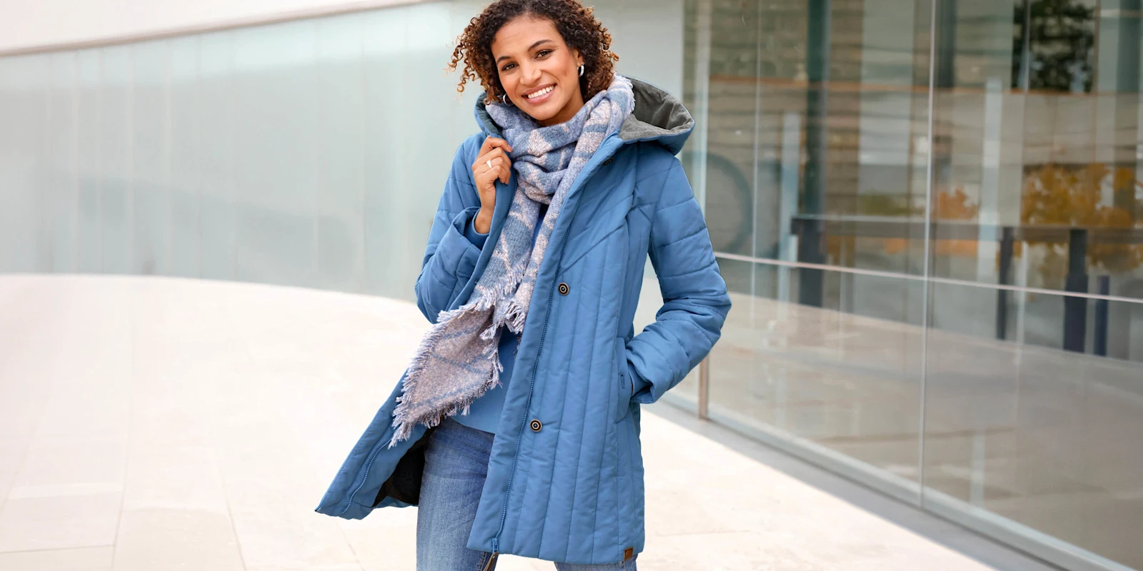 Een model draagt een blauwe gewatteerde jas, jeans en een sjaal met patroon voor een gebouw.