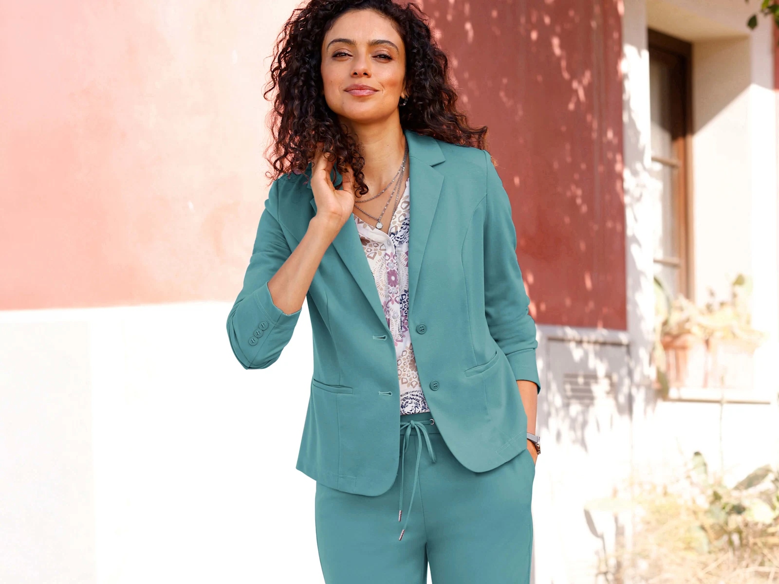 Ein Model trägt einen türkisfarbenen Anzug mit Blazer und Hose, kombiniert mit weißem T-Shirt und weißen Sneakern, steht draußen vor einem Haus.