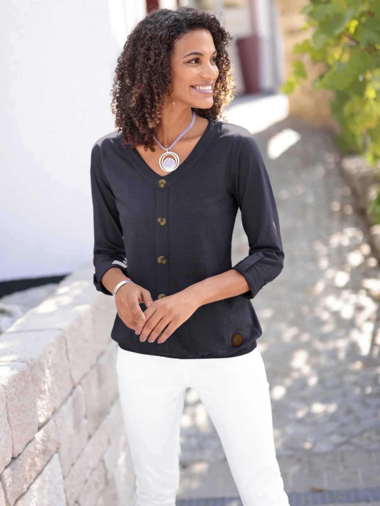 Frau trägt eine schwarze Bluse mit V-Ausschnitt und Knöpfen sowie weiße Hose, im Hintergrund eine helle Steinmauer und grüne Pflanzen.