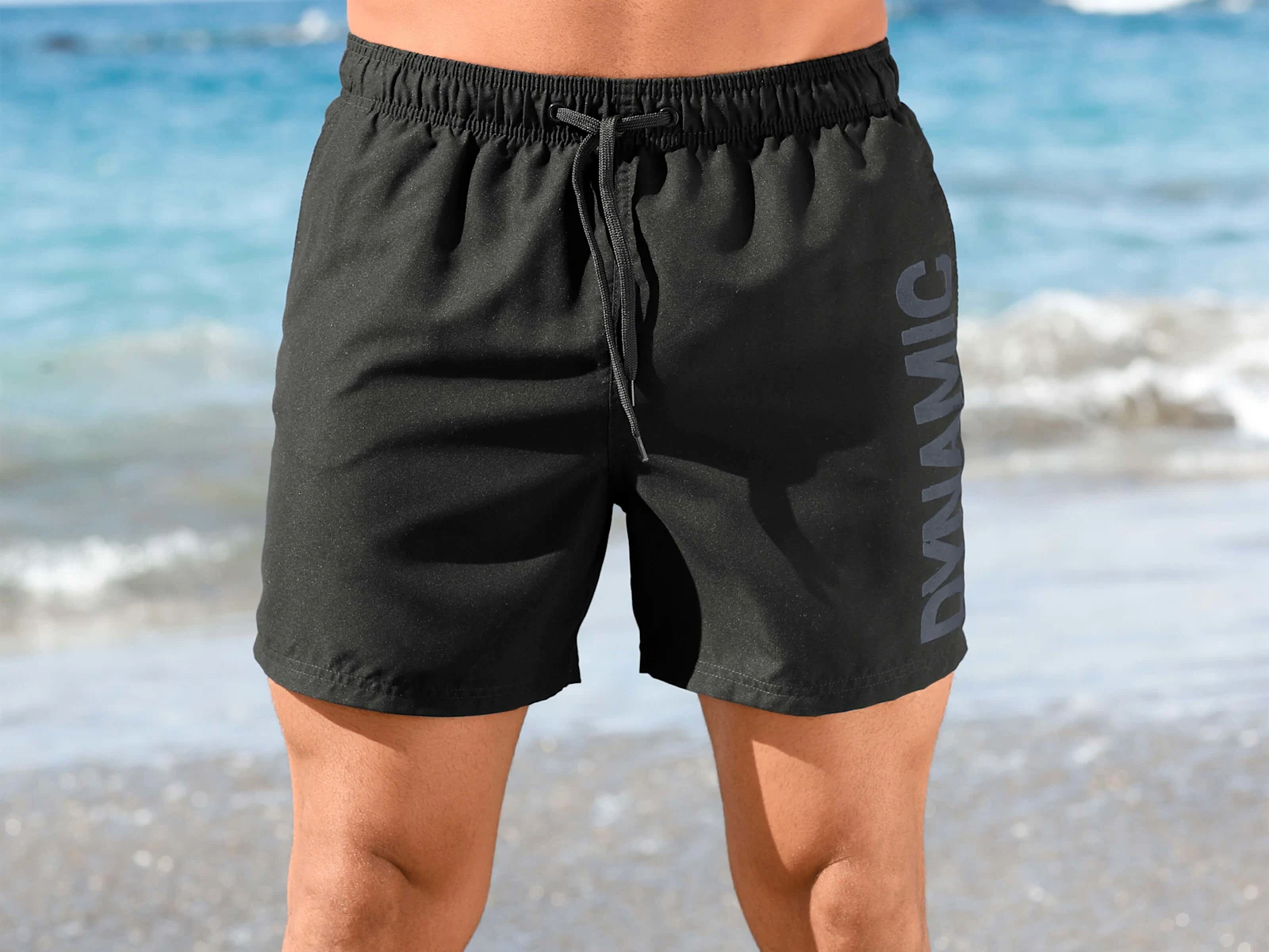 Ein männliches Model steht am Strand und trägt eine schwarze Badehose mit Kordelzug und seitlichem Schriftzug 'DYNAMIC'.