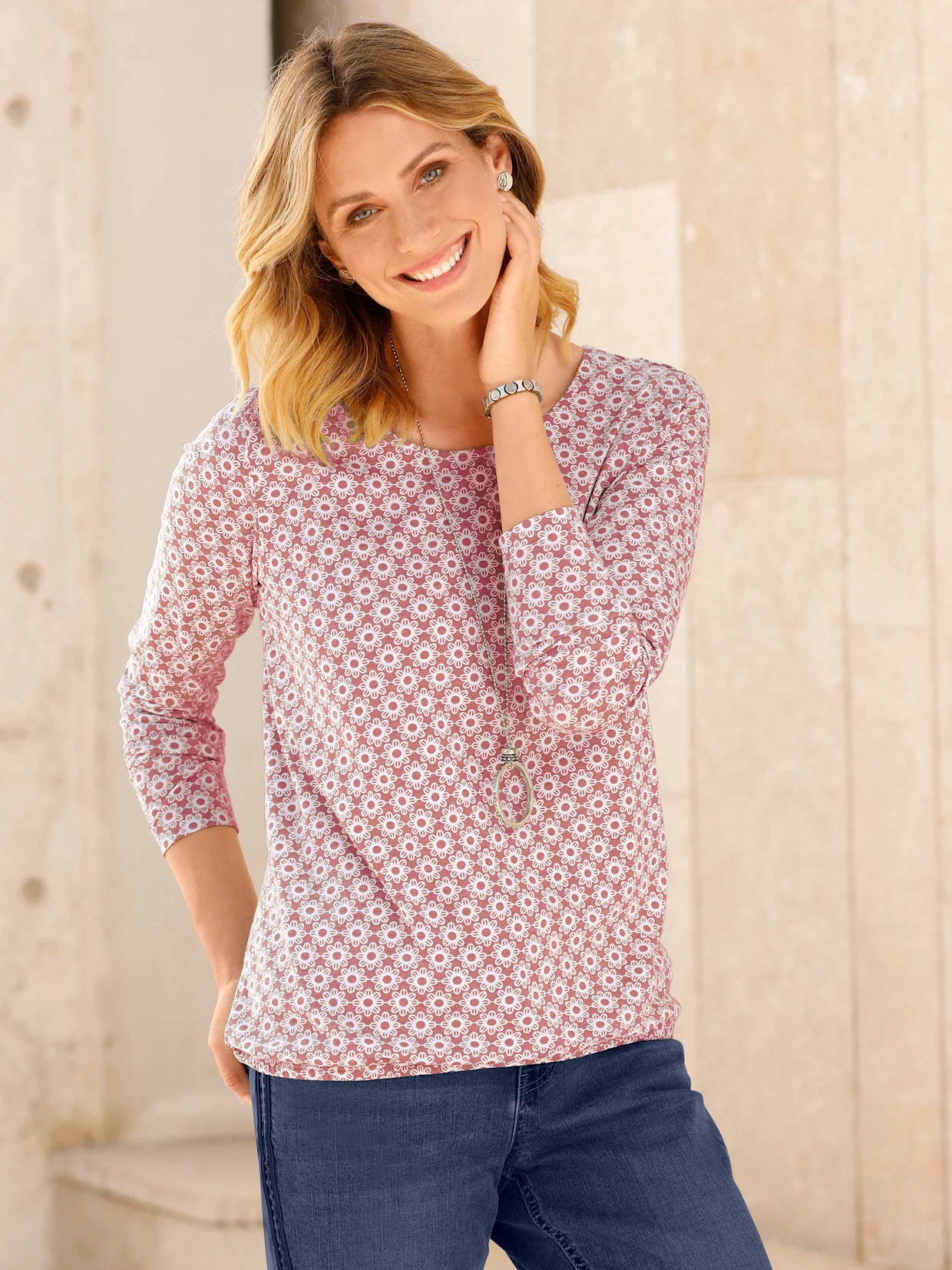 Vrouw draagt een roze shirt met bloemenpatroon, gecombineerd met een blauwe jeans en een lange halsketting.