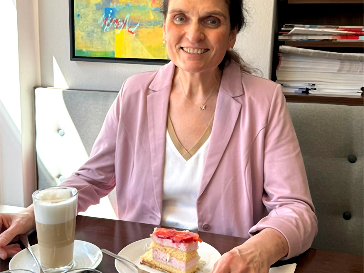 Eine Frau sitzt an einem Tisch mit einer Tasse Kaffee und einem Stück Kuchen. Sie trägt einen rosa Blazer, eine weiße Bluse und eine Kette. Im Hintergrund sind eine Wand und einige Unterlagen sichtbar. Eine Frau sitzt an einem Tisch mit einer Tasse Kaffee und einem Stück Kuchen. Sie trägt einen rosa Blazer, eine weiße Bluse und eine Kette. Im Hintergrund sind eine Wand und einige Unterlagen sichtbar.