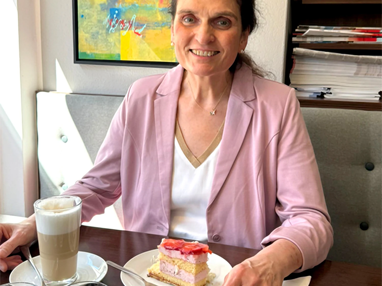 Eine Frau sitzt an einem Tisch mit einer Tasse Kaffee und einem Stück Kuchen. Sie trägt einen rosa Blazer, eine weiße Bluse und eine Kette. Im Hintergrund sind eine Wand und einige Unterlagen sichtbar. Eine Frau sitzt an einem Tisch mit einer Tasse Kaffee und einem Stück Kuchen. Sie trägt einen rosa Blazer, eine weiße Bluse und eine Kette. Im Hintergrund sind eine Wand und einige Unterlagen sichtbar.