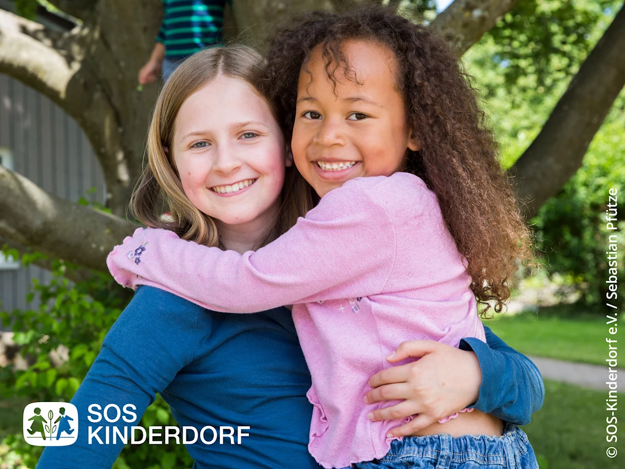 Zwei Kinder umarmen sich draußen vor einem Baum, fröhliche Atmosphäre im Grünen. Logo von SOS Kinderdorf unten links. Zwei Kinder umarmen sich draußen vor einem Baum, fröhliche Atmosphäre im Grünen. Logo von SOS Kinderdorf unten links.